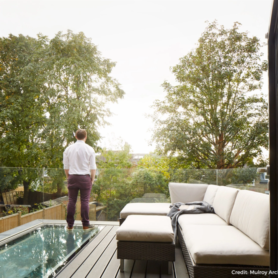 Ein Mann steht auf einer Veranda mit Blick auf Bäume und den Himmel. Er trägt ein weißes Hemd und lila Hosen. Eine gemütliche Couch mit Kissen ist im Vordergrund sichtbar.