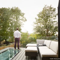 A man stands on a balcony, gazing across a landscape filled with trees. He is dressed in a white shirt and dark pants, with a sofa and a throw blanket beside him. The sky is bright, indicating a sunny day.
