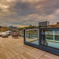 A spacious rooftop terrace featuring wooden flooring, outdoor seating arrangements, and glass railings. The skyline includes buildings and a cloudy sky in the background.