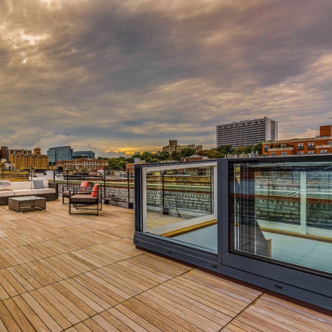 Terraza amplia con suelo de madera, muebles modernos y vistas de una ciudad al atardecer, con nubes grises en el cielo.