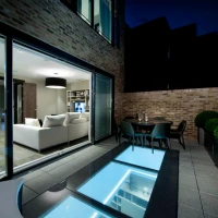 Moderne Terrasse bei Nacht mit einem eleganten Wohnraum im Hintergrund. Der Boden hat einen gläsernen Bereich, der das Licht reflektiert. Es gibt einen Esstisch mit Stühlen und gepflegte Pflanzen in Containern.