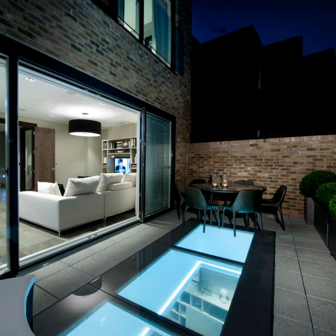 A modern patio features large glass doors leading to a stylish living room. The outdoor space has a sleek dining area with a round table and planters. A unique glass floor panel illuminates the area, adding a contemporary touch. Its night time, and the ambiance is cozy with soft lighting.