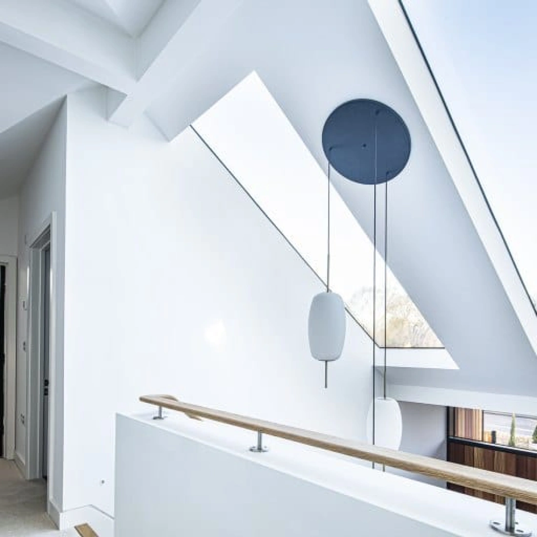 A modern, well-lit hallway with a sloped ceiling and large skylights. A sleek, minimalistic hanging light fixture is suspended from the ceiling. The walls are painted white, and the railing is made of wood.