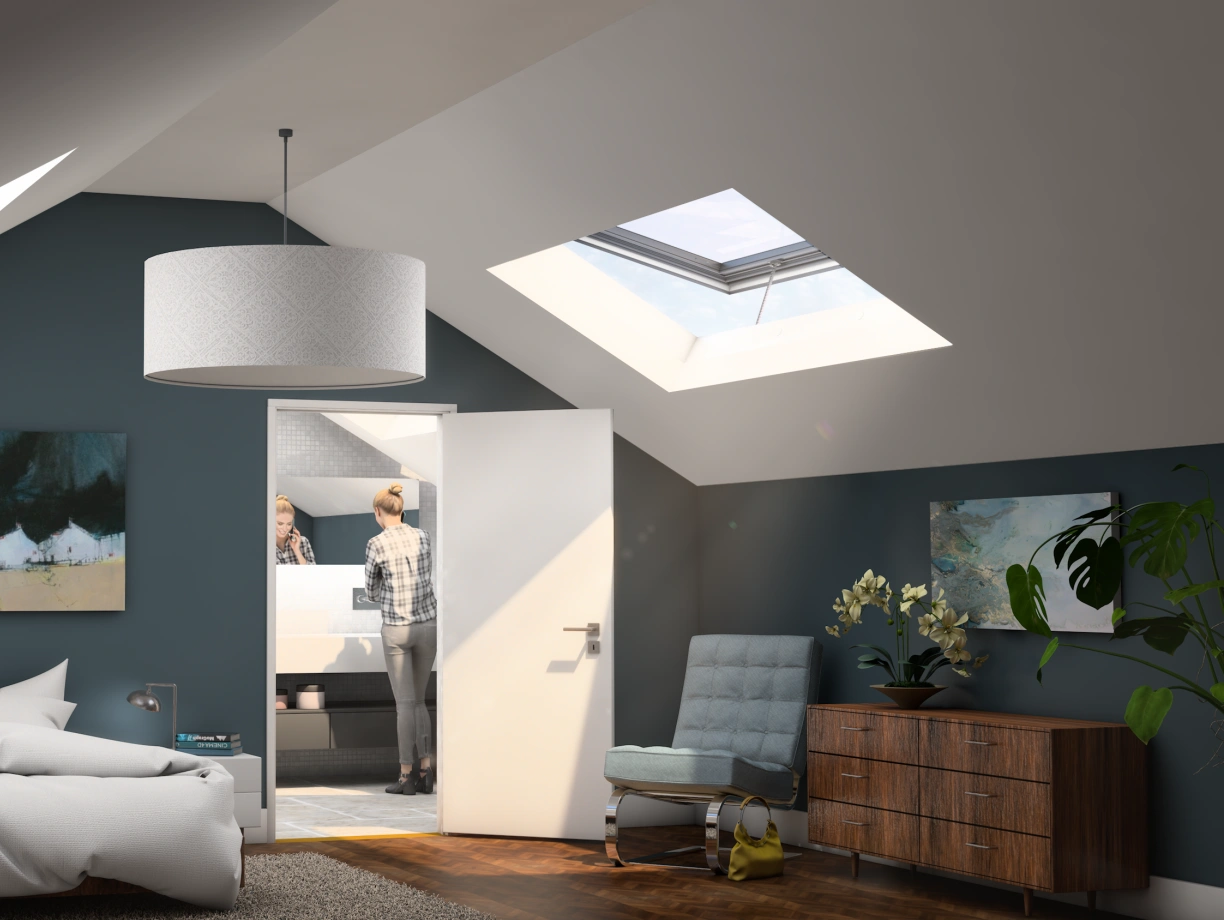 Habitación moderna con paredes azul oscuro, cama deshecha, dos cuadros grandes y un mueble de madera. Una puerta abierta muestra un baño y una persona de espaldas frente al espejo. Ventanas de techo permiten la entrada de luz natural.