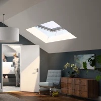 Bright and modern attic bedroom featuring a bed with white bedding, a stylish bedside lamp, and artwork on the walls. A woman is seen in the adjoining bathroom, while large skylights let in natural light. A wooden dresser and a contemporary chair complete the cozy atmosphere.
