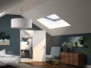 A modern attic bedroom featuring a large bed with crumpled white linens, a sleek bedside lamp, and a wooden dresser. Two skylights illuminate the space, and a person is visible in the bathroom through an open door. Abstract art hangs on the walls, and a potted plant adds a touch of greenery.