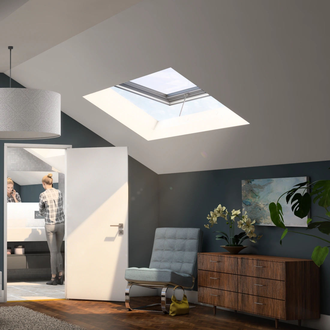 A modern attic bedroom featuring a large bed with crumpled white linens, a sleek bedside lamp, and a wooden dresser. Two skylights illuminate the space, and a person is visible in the bathroom through an open door. Abstract art hangs on the walls, and a potted plant adds a touch of greenery.