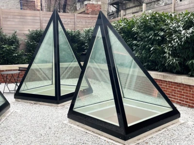 Two modern glass and black-frame pyramidal structures are placed on a gravel surface in a courtyard, surrounded by low greenery and brick walls. An apartment building with fire escapes is visible in the background.