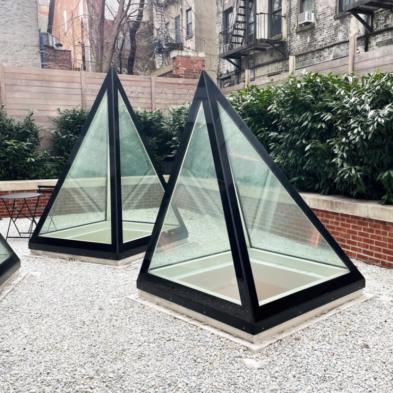 eux structures en forme de pyramide en verre, avec des bases noires, se trouvent dans une cour pavée de galets. En arrière-plan, on aperçoit des murs en briques et des fenêtres d’immeubles.