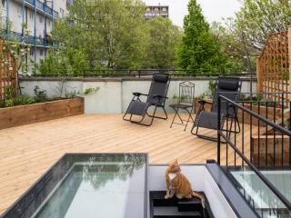 A rooftop garden featuring wooden decking, two black lounge chairs, a small table, and a cat sitting near a set of stairs that lead to a lower level. Lush greenery surrounds the area with buildings in the background.