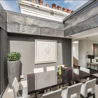 A modern dining area features a sleek dark table surrounded by white chairs. A large abstract artwork hangs on the gray wall, and greenery adds a touch of nature. Natural light streams in from a skylight above, illuminating the space.