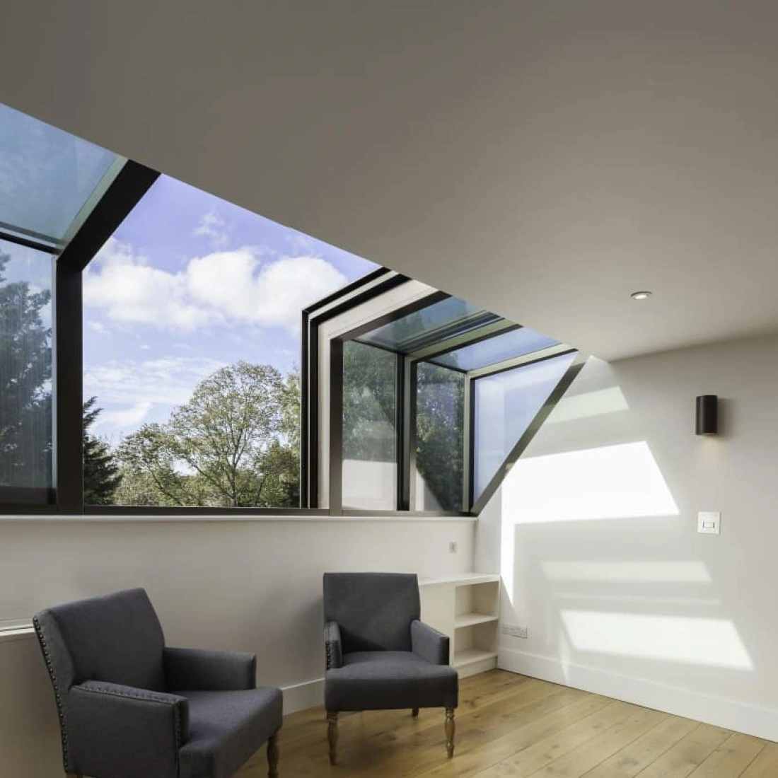 Two blue upholstered armchairs sit on a wooden floor in a bright room with large windows. The angular window design allows natural light to fill the space, and trees are visible outside. A small shelf is located beside the chairs.