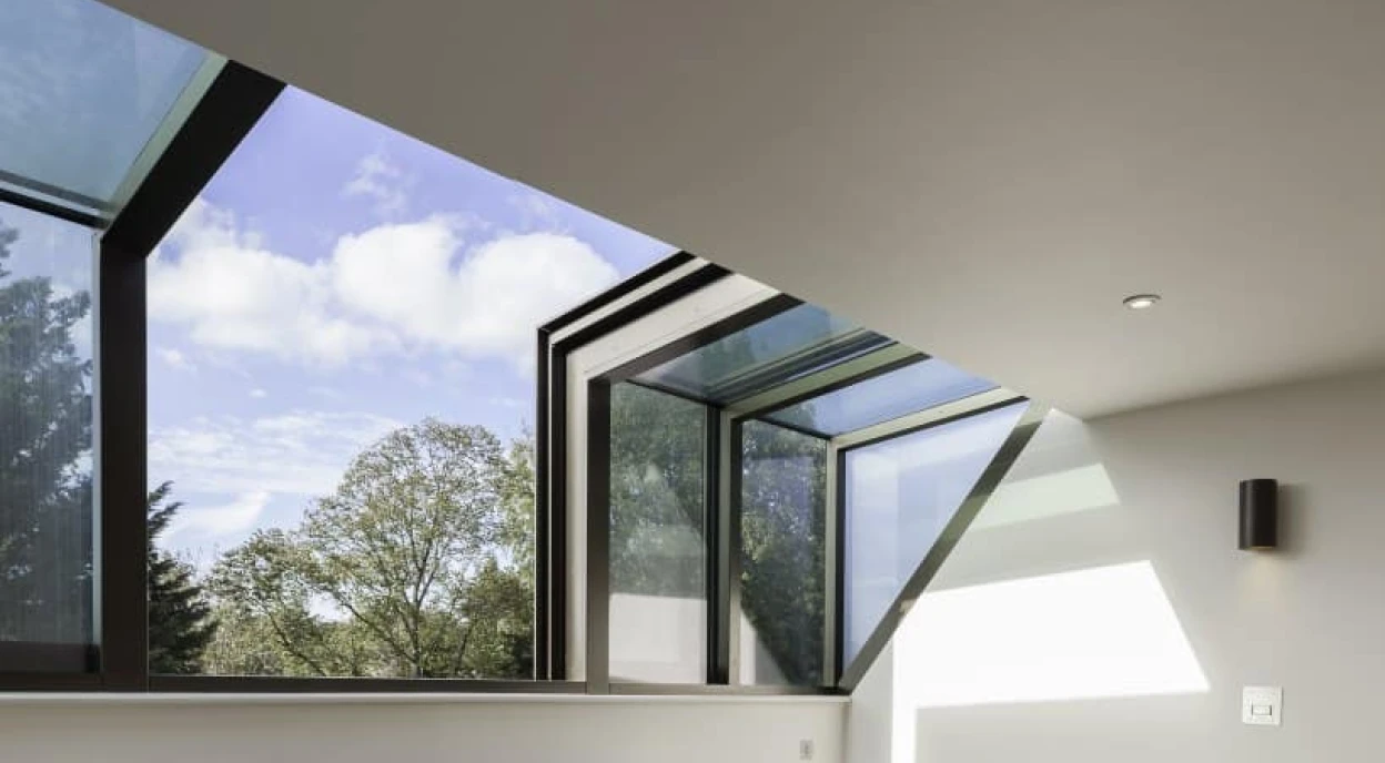 Two blue armchairs with wooden legs sit on a light wooden floor in a bright room. Large windows with a modern design allow natural light to fill the space, showcasing a view of trees and a partly cloudy sky. A built-in shelf is positioned against the wall.