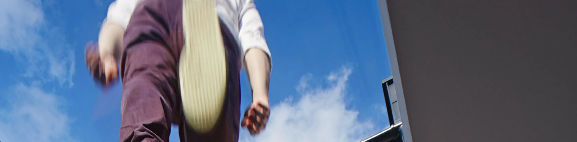 Eine Person springt von einer erhöhten Position auf ein Gebäude mit Backsteinfassade. Der Blick ist von unten durch ein Glasdach gerichtet, das den Himmel sichtbar macht.