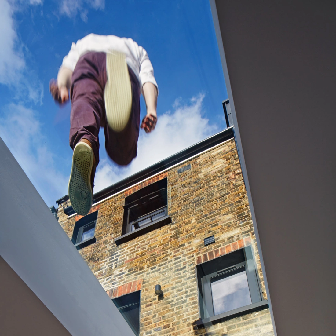 A person is seen walking on a glass skylight, with their feet visible above a brick building. The blue sky peeks through the skylight.