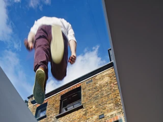 Una persona con pantalones oscuros y zapatos claros parece estar caminando sobre un tragaluz, con una vista del cielo azul y nubes. La pared de ladrillo expuesta de una casa se ve detrás.