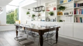 A modern dining area with a large wooden table and transparent chairs. White shelves filled with books, plants, and decorative items line the walls. A skylight allows natural light to flood in, creating a bright and airy atmosphere.